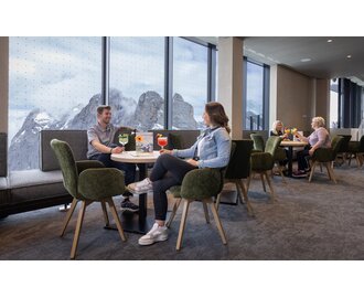 People enjoying drinks at tables with view of snow-covered mountains through panorama windows. | © Harald Steiner