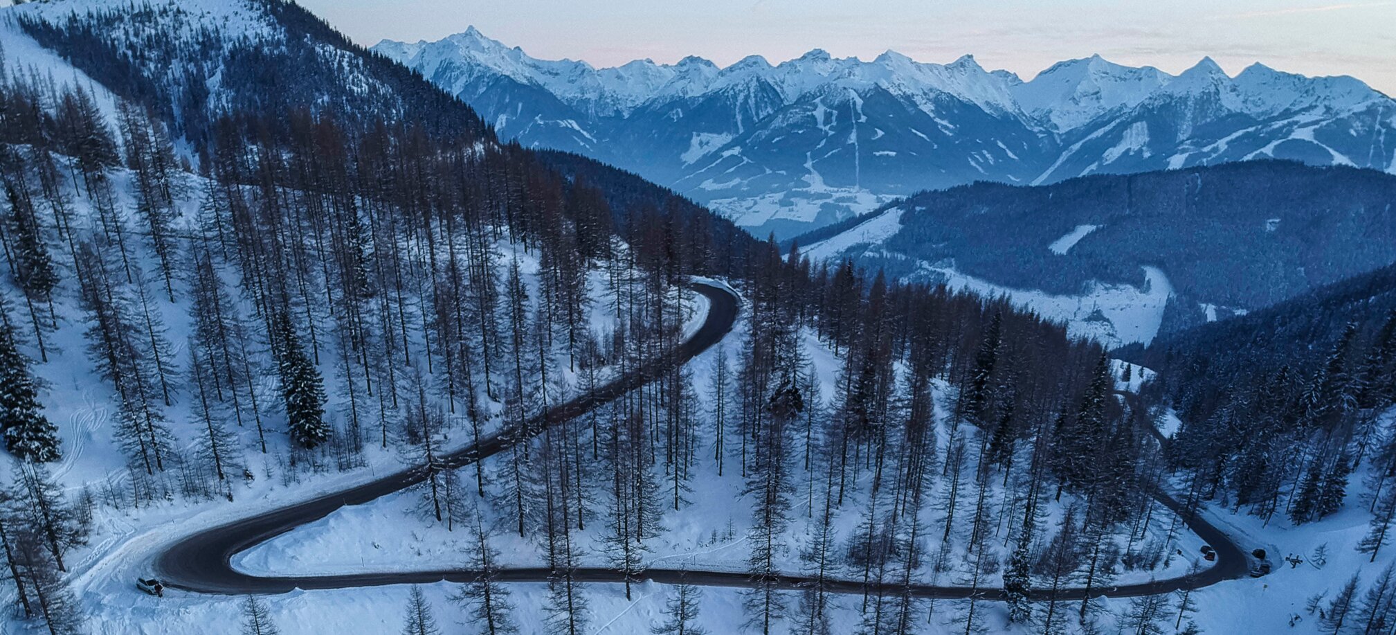 Snowy serpentine road through coniferous forest with view of snow-covered mountains. | © David McConaghy