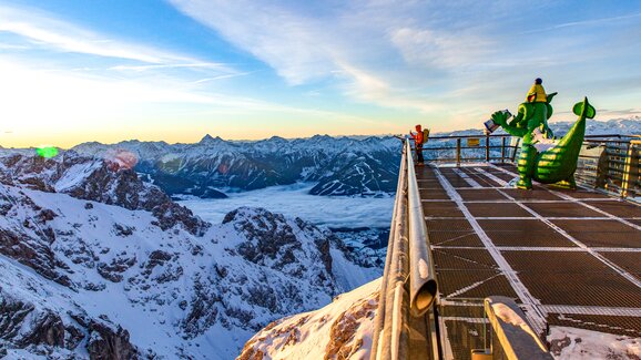 Snow-covered mountains, man on viewing platform, and green dragon figure at Dachstein. | © Renè Eduard Perhab