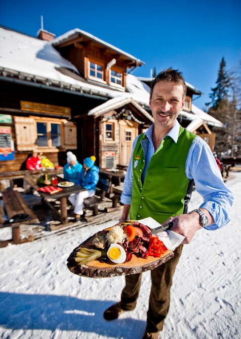 Man with snack board in front of wooden alpine hut, people in background | © Tom Lamm