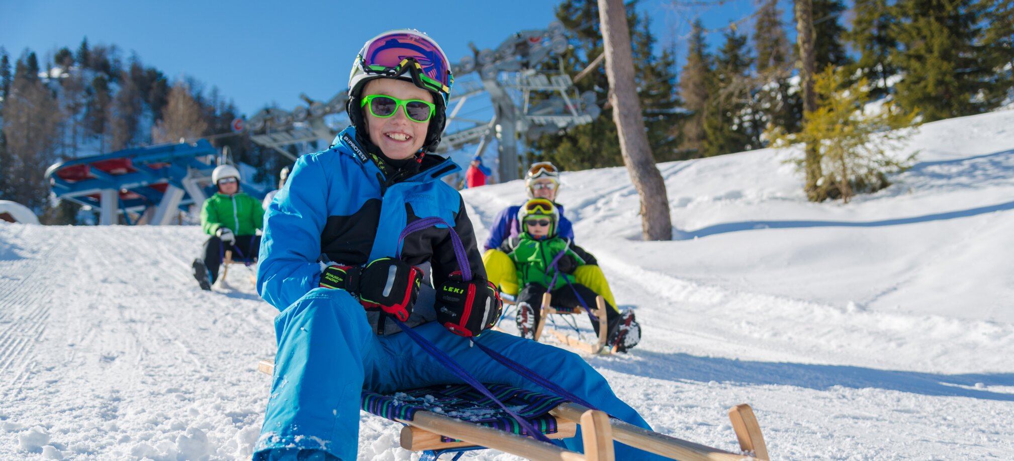 Smiling boy with sunglasses rides sled in snow, others follow behind. | © Christine Höflehner