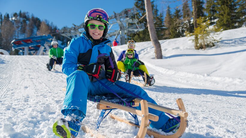 Smiling boy with sunglasses rides sled in snow, others follow behind. | © Christine Höflehner