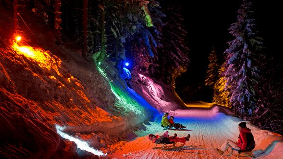 Colorful lights illuminate night sledding trail with riders in the snow. | © Herbert Raffalt