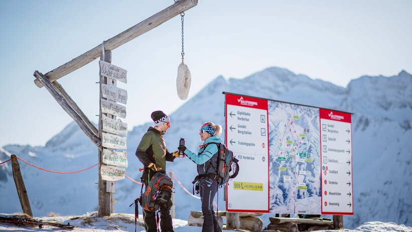 Two ski tourers stand at a weather station with wooden sign and panorama map. | © Christoph Huber
