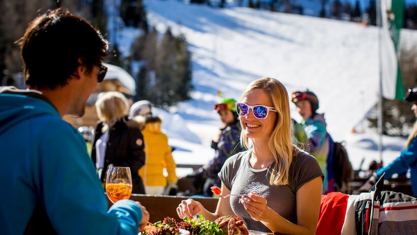 Woman with sunglasses enjoying food at the ski hut in sunshine, skiers in the background. | © Tom Lamm