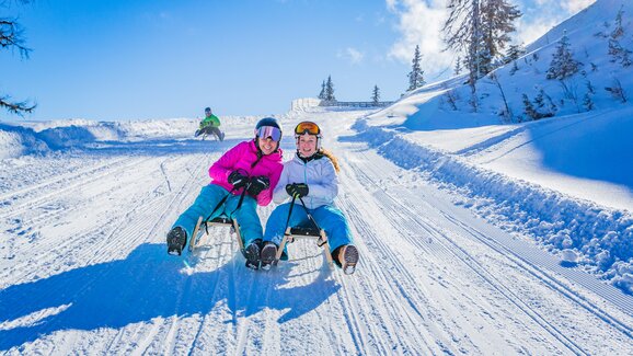 Two women sledding and laughing on snowy toboggan run with alpine backdrop. | © René Eduard Perhab