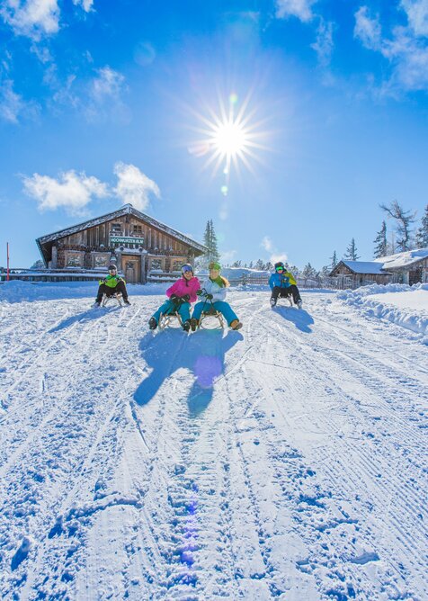 Sledders in front of Hochwurzenalm on snowy run with bright sunshine. | © René Eduard Perhab