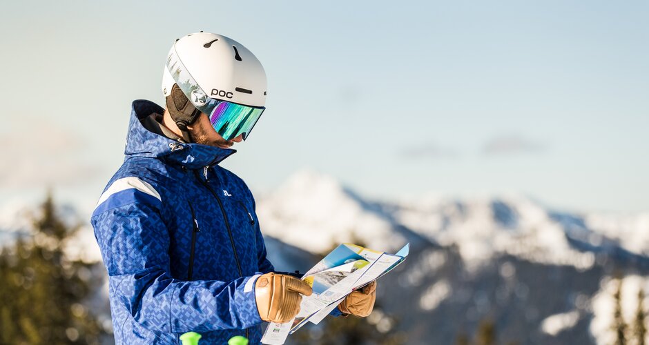 Skier in blue jacket and white helmet looks at ski map with alpine panorama. | © Dominik Steiner
