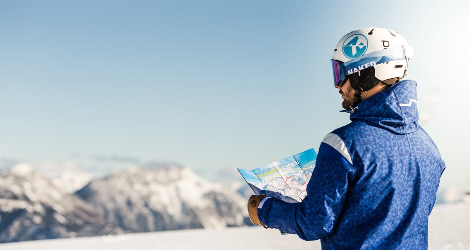 Skier in blue jacket with helmet looks at ski map in front of snowy mountains. | © Dominik Steiner
