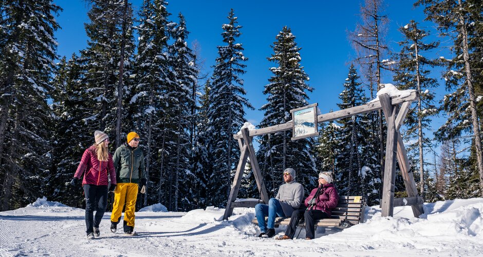 Winter hikers and seated guests on a wooden bench in front of snowy trees. | © Gerald Oberreiter