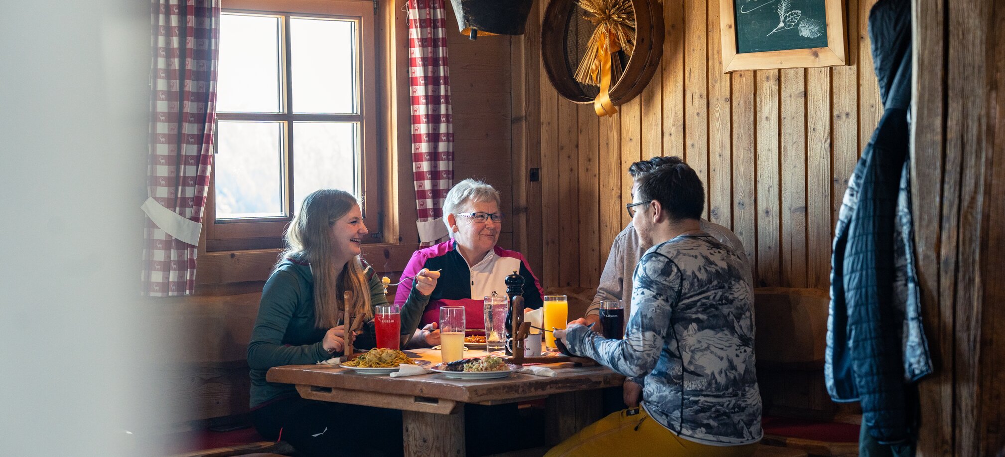 Four people laugh while eating in a wooden hut, drinks on the table. | © Gerald Oberreiter
