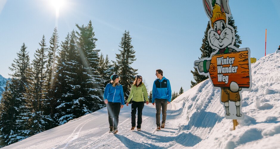 Three hikers walk along the winter hiking trail with Hopsi mascot in the background. | © Christine Höflehner