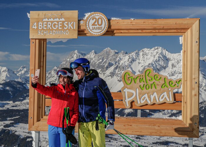 Two skiers take a selfie in front of a wooden frame with snowy mountains. | © Herbert Raffalt