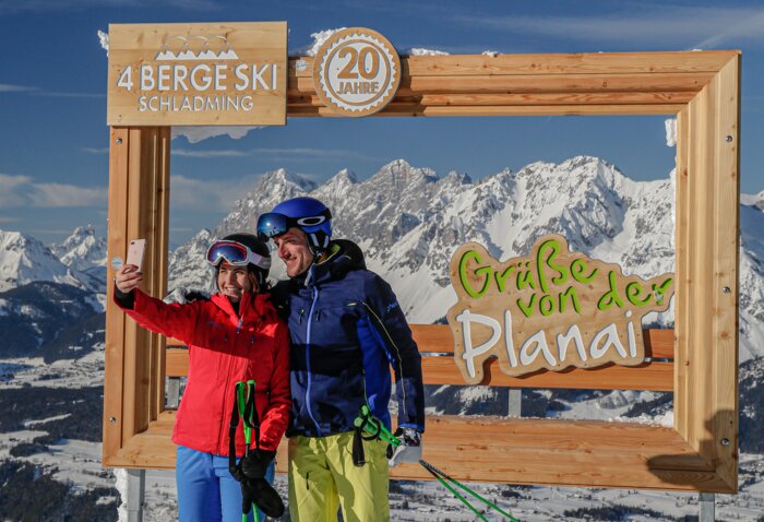 Zwei Skifahrer machen ein Selfie vor einem Holzrahmen mit verschneiten Bergen. | © Herbert Raffalt