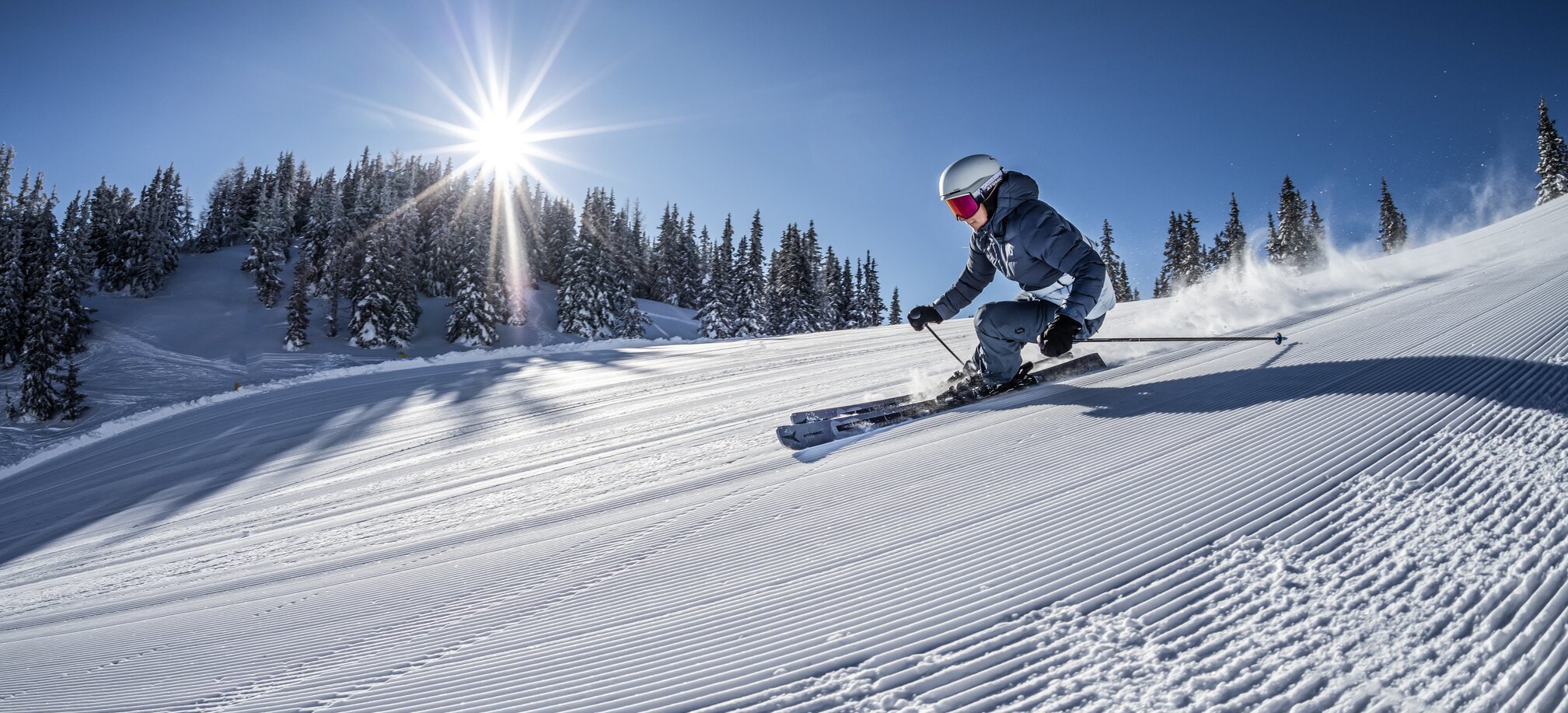 Skier carves a groomed slope under the sun with snowy trees in the background. | © Mirja Geh