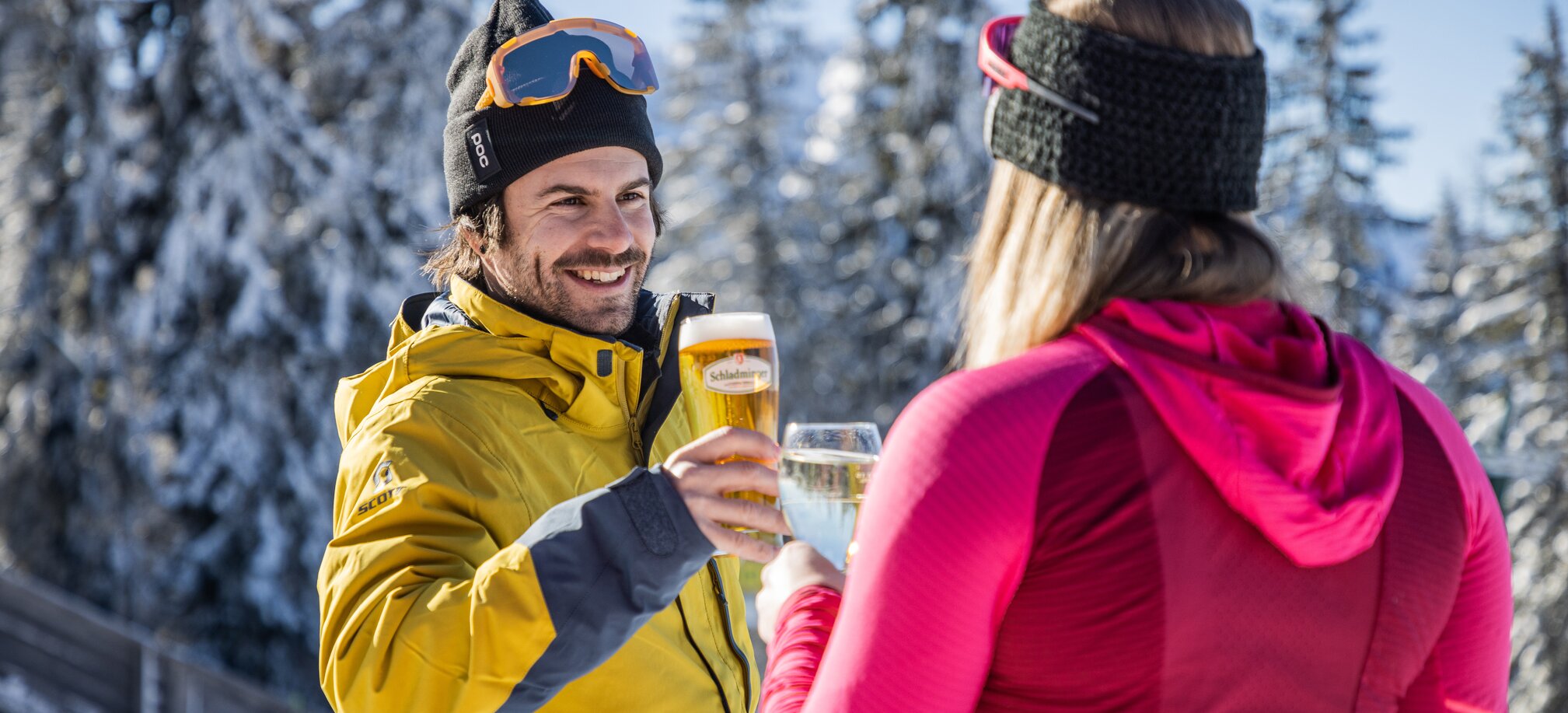 Two people clinking beer and wine glasses in front of a snowy winter landscape. | © Mirja Geh