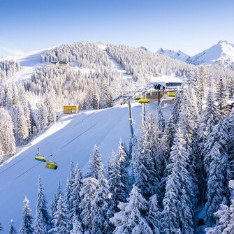 Aerial view of Planai with snowy trees, ski slopes, and a yellow cable car against a blue sky. | © Josh Absenger