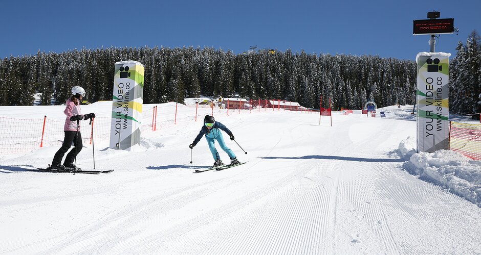 Skier in a blue suit reaches the finish line of the Skimovie racecourse on Planai, while a companion watches. | © Herbert Raffalt