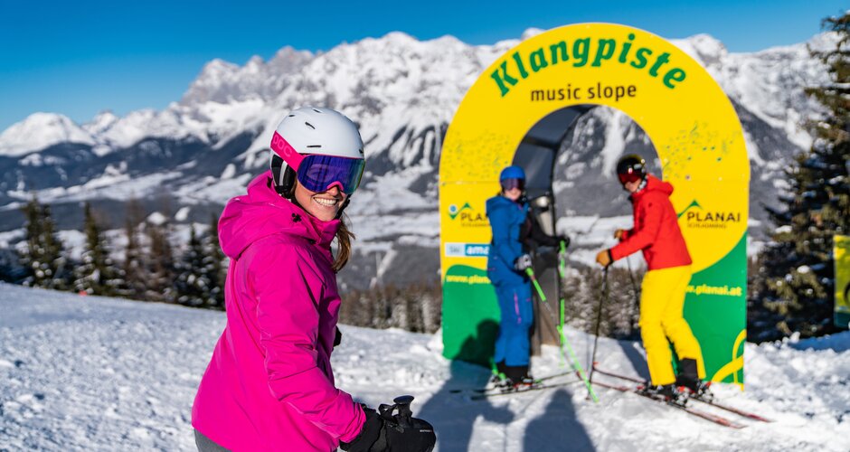Three skiers at yellow gate of music slope on Planai with view of Dachstein | © Tom Lamm