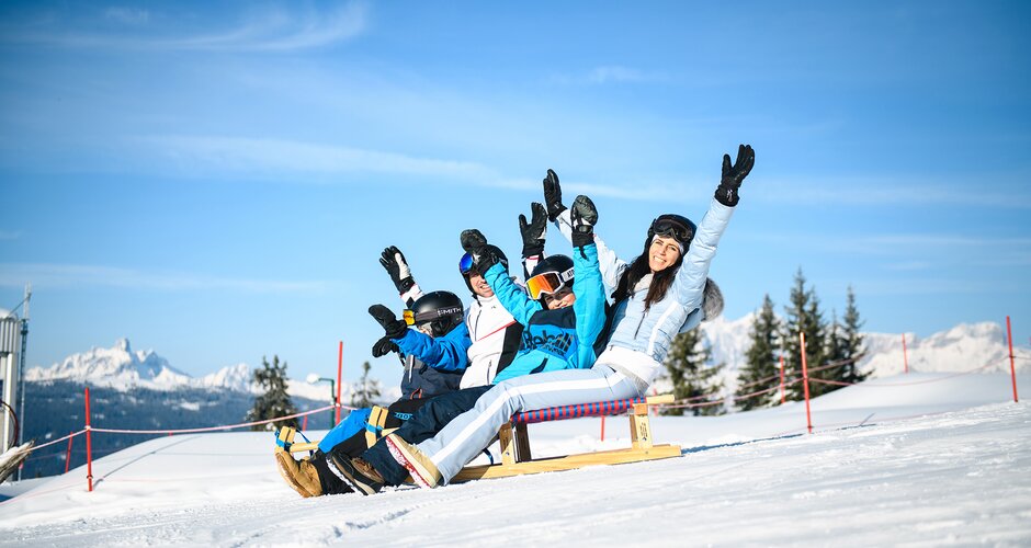 Four people laughing while sledding on the snowy Königslehen toboggan run.