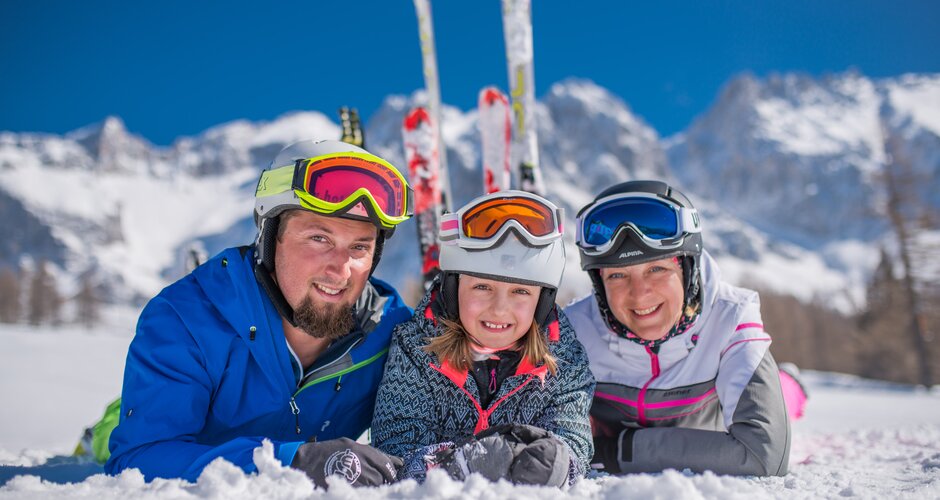 Family with ski goggles lying in the snow, smiling in front of snowy mountains. | © Christine Höflehner