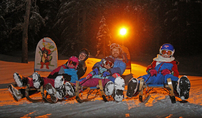 Family night sledding with helmets and headlamps on snowy track.