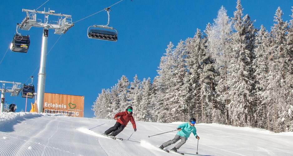 Two skiers ski down a freshly groomed slope and the mountain station of the combined lift can be seen above