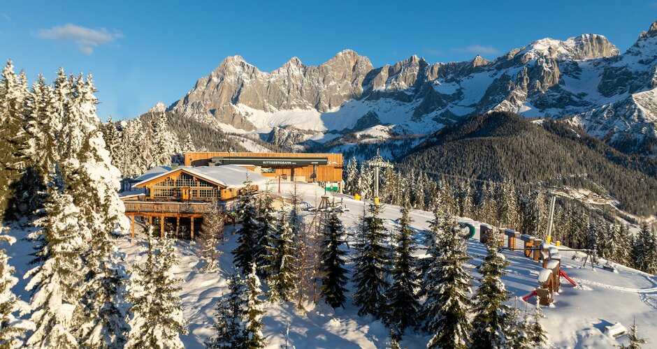 Bergstation der Rittisbergbahn mit Hütte, Spielplatz und Dachstein im Sonnenlicht.