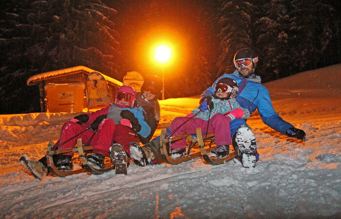 Two adults and two kids tobogganing at night on Rittisberg under floodlight