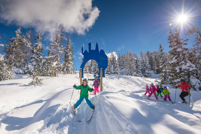 Children skiing through a castle-shaped arch surrounded by snowy trees. | © Lorenz Masser