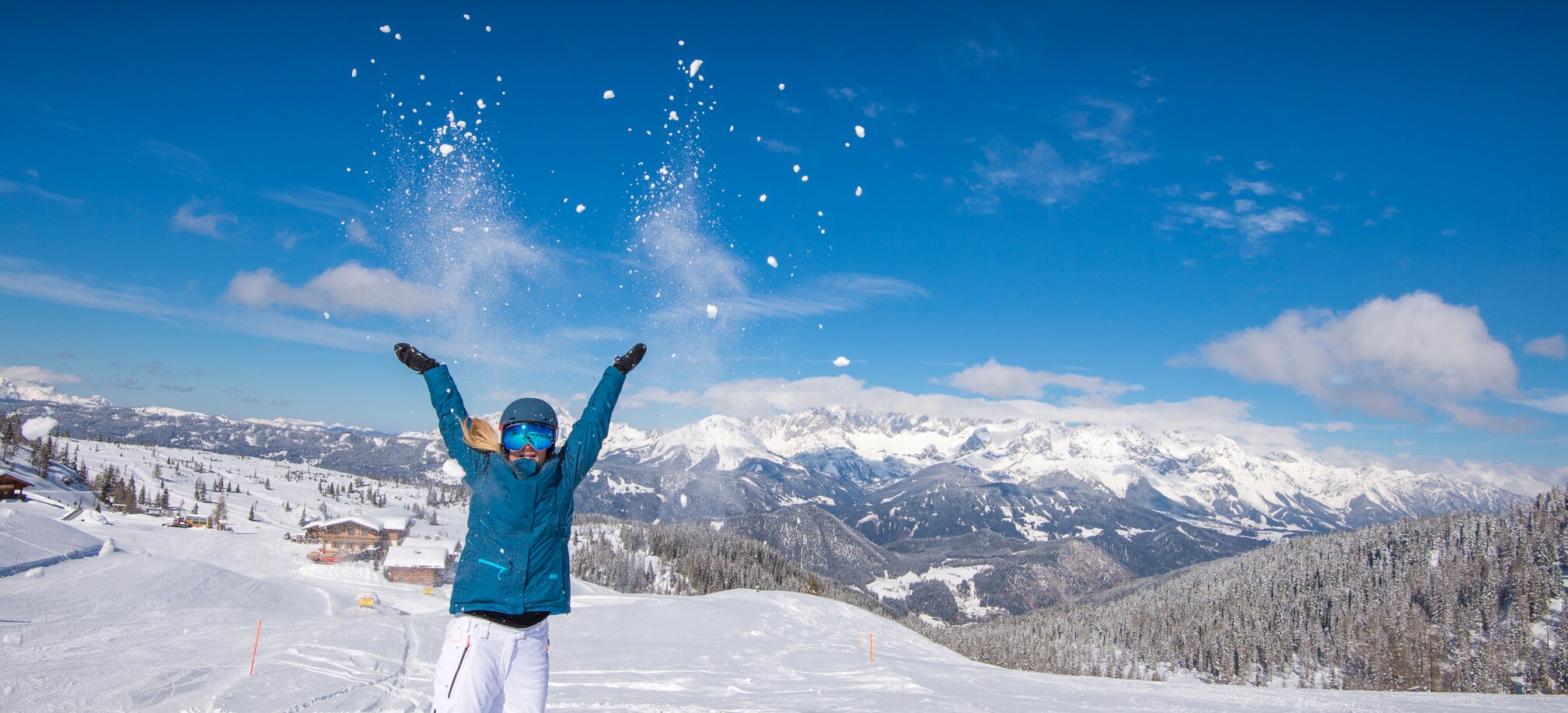 Woman throws snow in the air, smiling, with mountains and snowy valley in background. | © Lorenz Masser