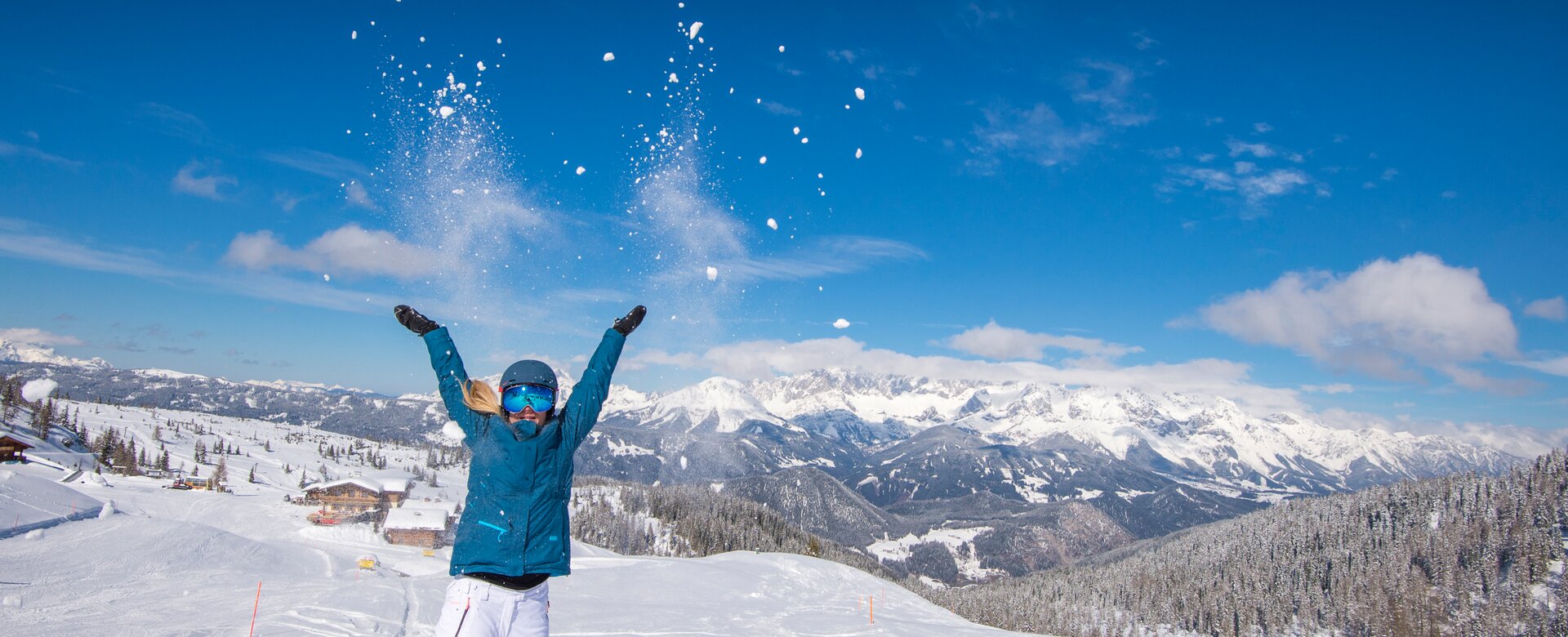 Woman throws snow in the air, smiling, with mountains and snowy valley in background. | © Lorenz Masser