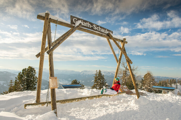 Person in red jacket on a wooden swing with alpine mountain view in the background. | © Lorenz Masser