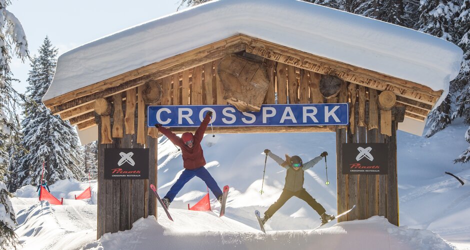 Two skiers jumping at the same time under a wooden "Crosspark" sign at Reiteralm. | © Lorenz Masser