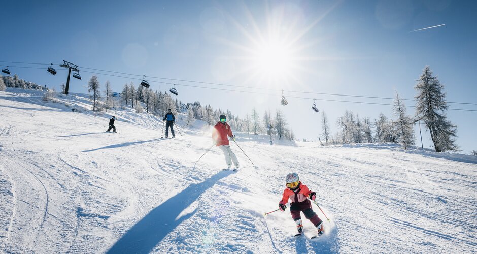 Child and adults skiing on a sunny slope, with chairlift and snowy trees in the background | © Shuttleberg