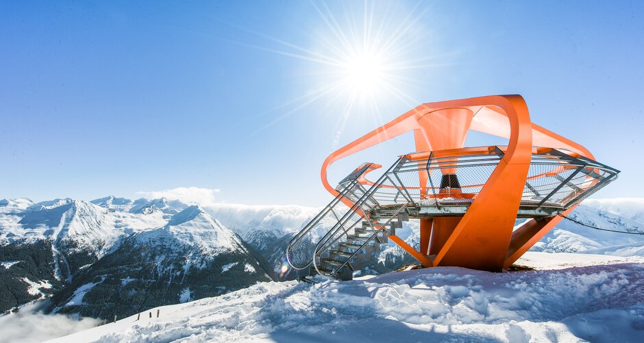 An orange viewing platform is surrounded by snow and a snow-covered mountain panorama | © Gasteiner Bergbahnen AG, Wolkersdorfer