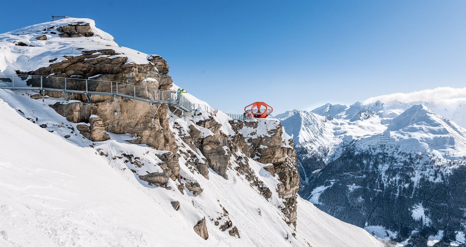 A metal path is fixed to a rock face with snow all around and the path leads to an orange viewing platform | © Gasteiner Bergbahnen AG, Wolkersdorfer