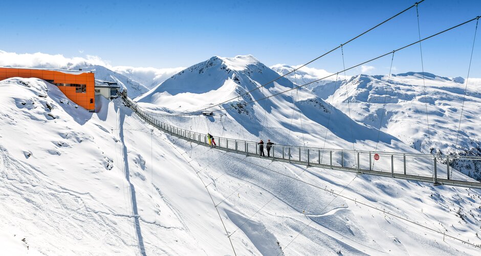 Suspension bridge spans a snow-covered precipice and at one end is an orange mountain station of a cable car | © Gasteiner Bergbahnen AG, Wolkersdorfer
