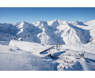  The top station of a cable car from above, with skiers next to it, surrounded by snow-covered mountains.