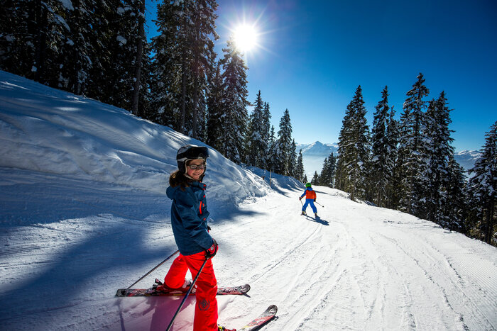 two children ride along a ski trail in a plough and one smiles into the camera