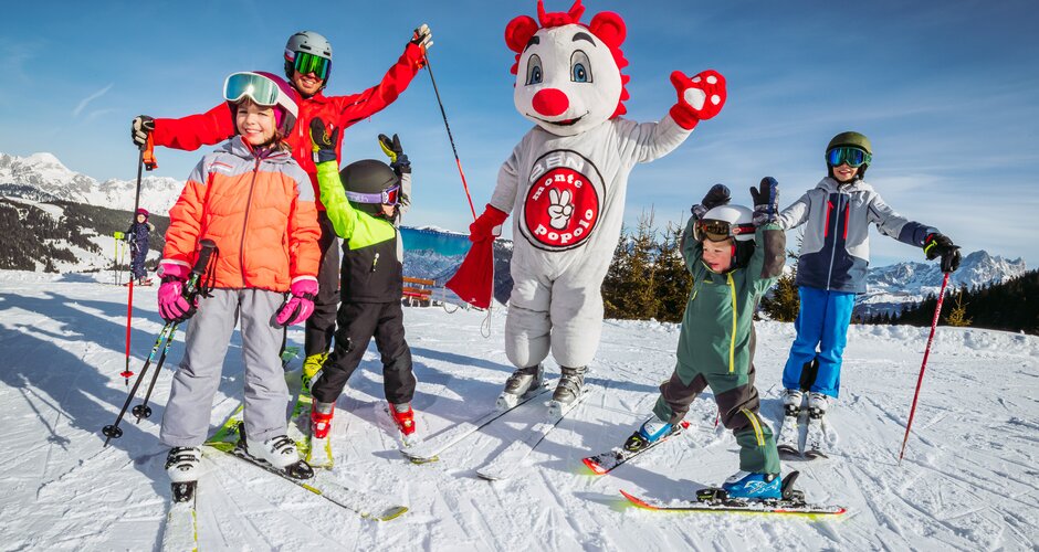 An adult and four children wave at the camera, with the Monte Popolo mascot in the middle. 