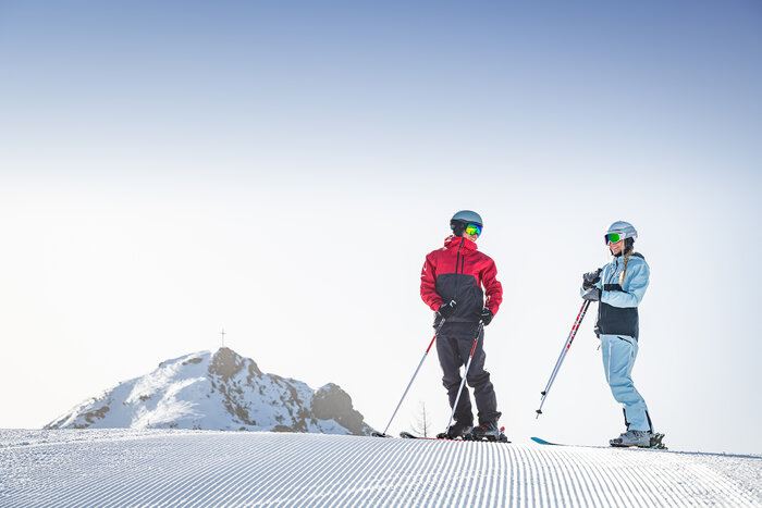 A female skier in a light blue ski suit and a male skier in a grey-red ski suit are standing smiling on a freshly prepared piste | © Snow Space Salzburg