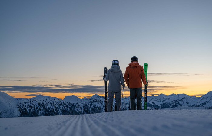 Two skiers hold their skis in their hands and look out at the sunrise