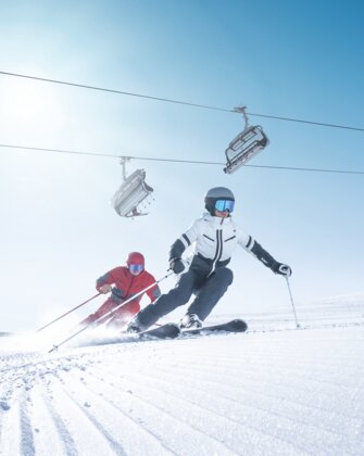 Zwei Skifahrer carven synchron auf frisch präparierter Piste bei Sonnenschein und Liftbetrieb