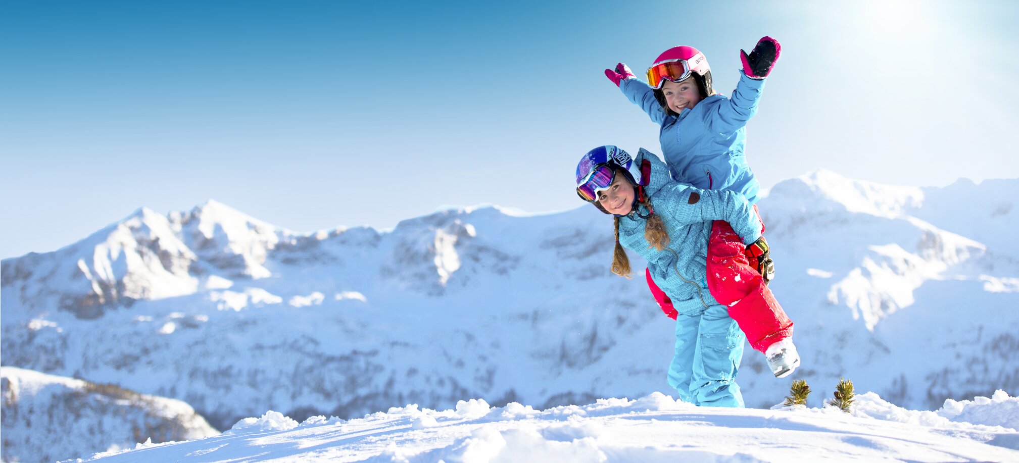 Two children in colorful ski outfits laugh and play in the snow before snowy peaks