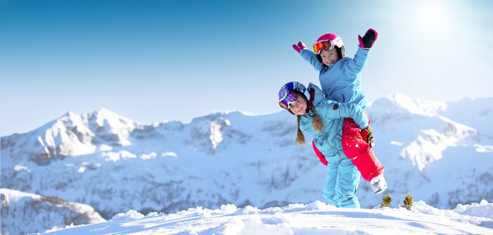 Two children in colorful ski outfits laugh and play in the snow before snowy peaks