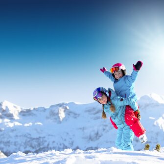 Two children in colorful ski outfits laugh and play in the snow before snowy peaks