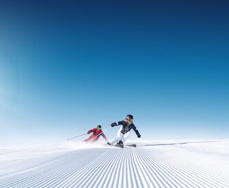 Two skiers carving dynamically on a freshly groomed slope under a clear blue sky