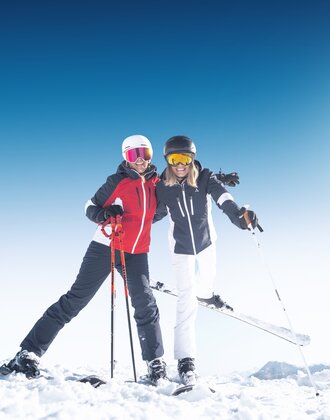 Two female skiers smiling in the snow with poles and helmets in bright sun at Ski amadé