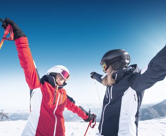 Two female skiers face each other with arms wide open, smiling in the snow and sunshine